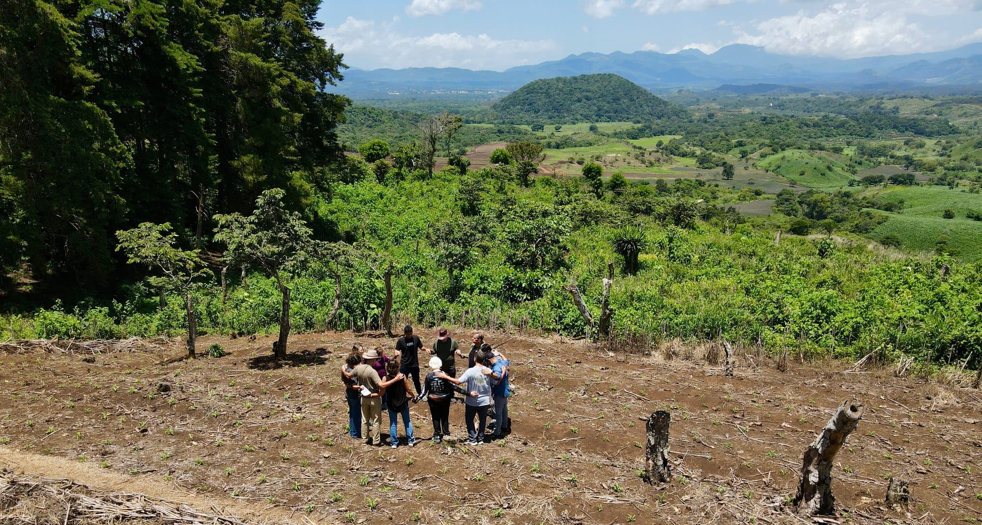 A group of people standing in a circle with their arms around each other on cleared hillside ground, with green hills and mountains beyond