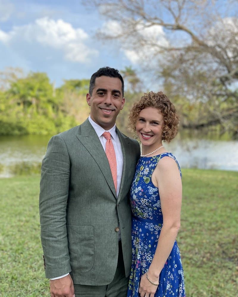 Miquel Gonzalez in a green suit standing outdoors beside a woman in a blue floral dress, with a lake and trees behind them